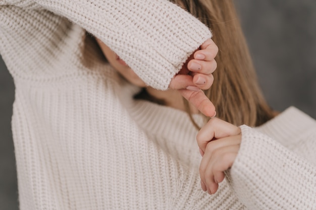 Una chica con un suéter ligero sobre una pared gris. Primer plano de las manos de las mujeres. La niña se cubre los ojos con las manos.