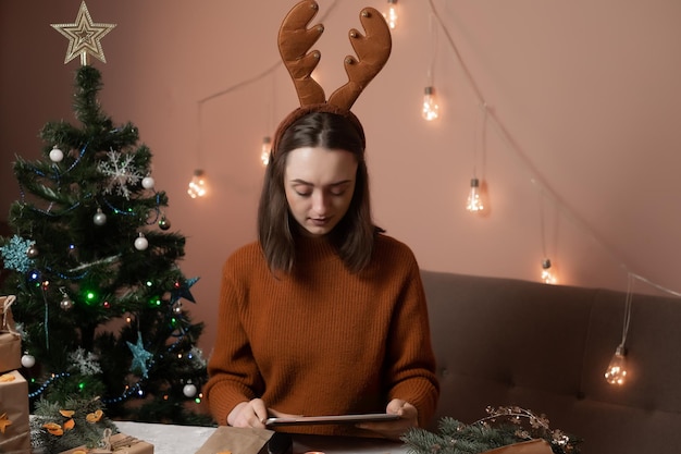 una chica con un suéter marrón sostiene una tableta y empaca regalos para el nuevo año