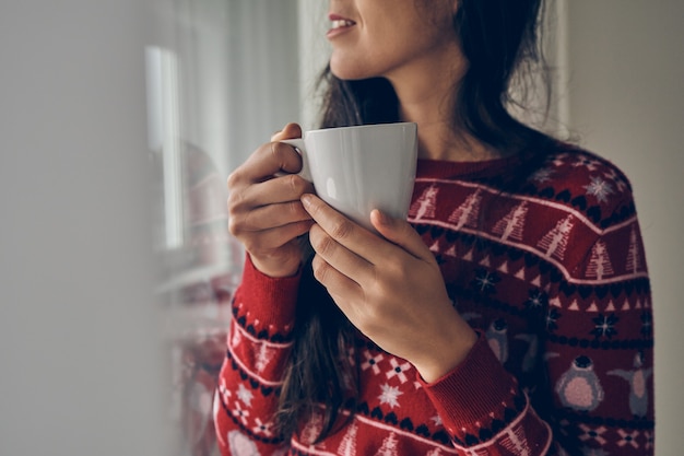 Chica con un suéter de Navidad sosteniendo una taza junto a la ventana