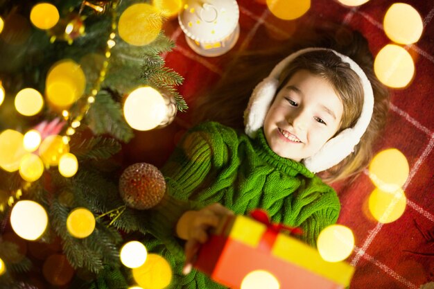 Chica con un suéter de punto cálido y auriculares de piel está acostada sobre una manta cerca del árbol de Navidad con una caja de regalo. Nochebuena, un sueño y un deseo. Año nuevo, ambiente festivo, guirnaldas y bokeh.