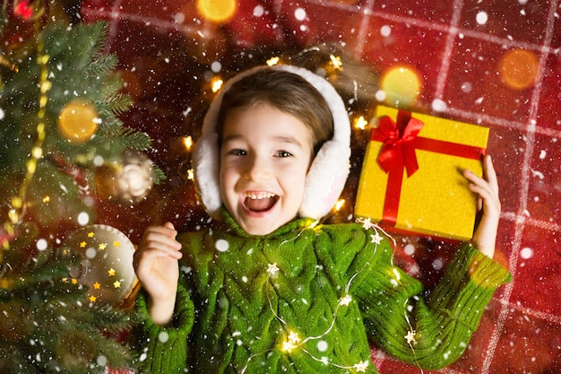 Chica con un suéter de punto cálido y auriculares de piel está acostada sobre una manta cerca del árbol de Navidad con una caja de regalo. Nochebuena, un sueño y un deseo. Año nuevo, ambiente festivo, guirnaldas y bokeh.
