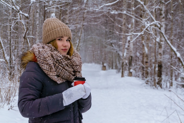 Chica en un suéter sosteniendo un vaso de papel con una bebida caliente en un parque de invierno