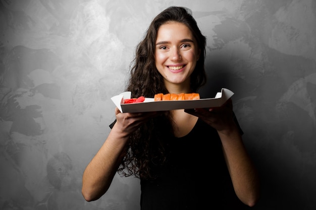 Chica sujetando filadelfia rollos en una caja de papel sobre fondo gris. Sushi, comida a domicilio.