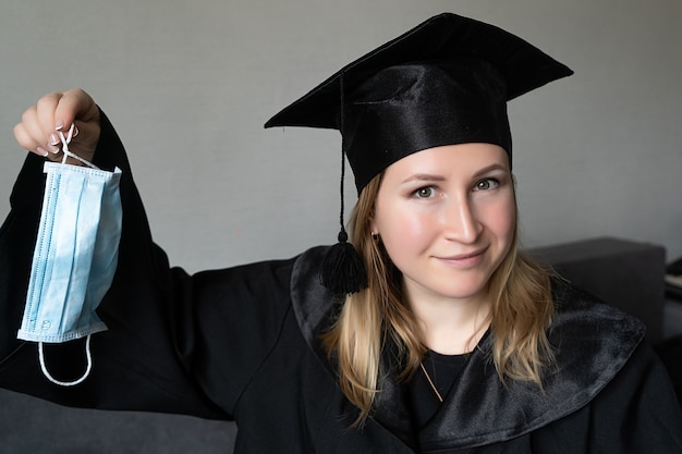 Chica sujetando máscara médica con sombrero de graduación sobre fondo gris