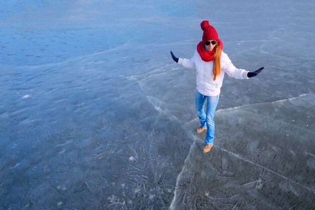 Chica en sunglasse, chaqueta blanca, gorra roja y bufanda roja manteniendo el equilibrio y divirtiéndose mientras camina sobre hielo en el lago congelado en invierno