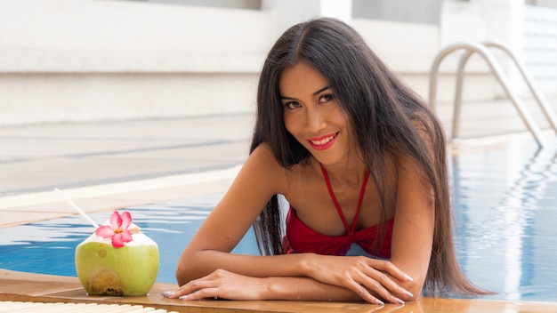 Foto chica tailandesa asiática sexy en bikini rojo de ropa interior de moda sentarse en la piscina con bebida de coco.