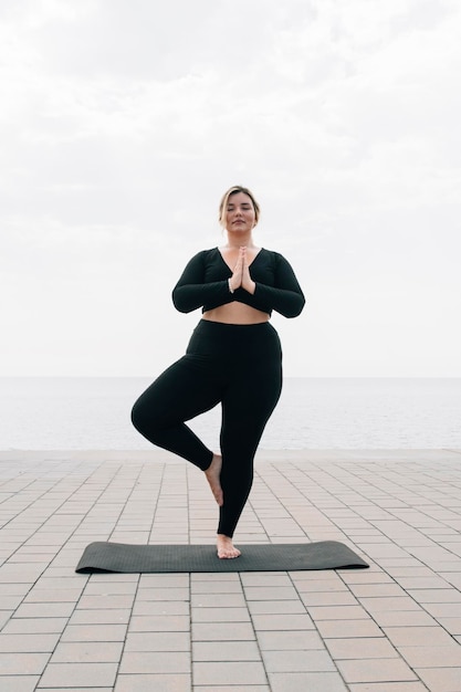 Chica de talla grande practicando yoga frente al mar en un día de verano
