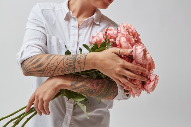 Chica con un tatuaje en sus manos, sosteniendo un ramo de hermosas rosas rosadas sobre un fondo gris. Día de la Madre