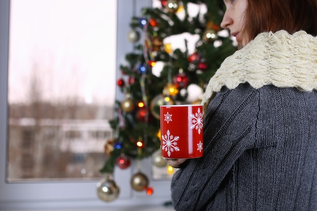 Chica con una taza de bebida caliente se encuentra frente a una ventana en la víspera de Año Nuevo