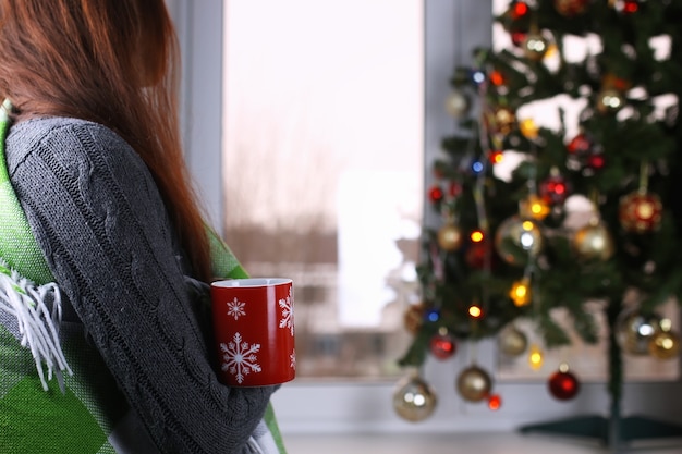 Chica con una taza de bebida caliente se encuentra frente a una ventana en la víspera de Año Nuevo