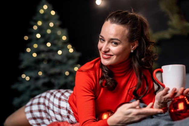 Chica con taza de bebida caliente relajante. Cosas que hacer antes de navidad. Mujer con taza y fondo de adornos navideños. Relájate y recarga energías.