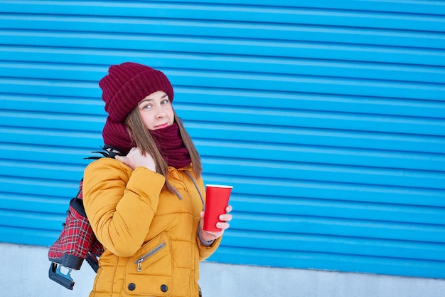Chica con una taza de café desechable y patines listos sobre su hombro sobre un fondo azul.