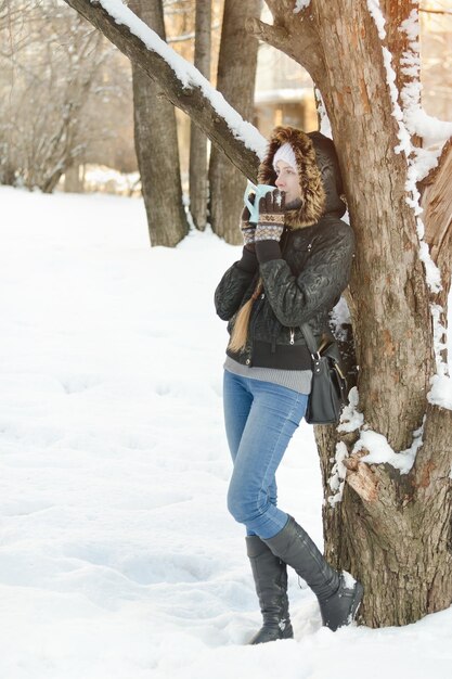 Chica con una taza de té apoyada contra un árbol y mirando directamente Invierno al aire libre