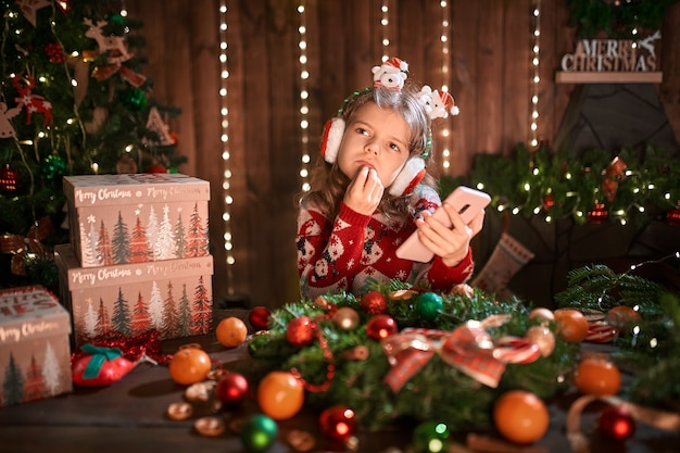 Chica con teléfono cerca del árbol de Navidad en el interior decorativo.