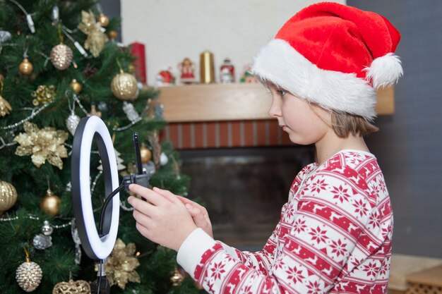 Chica con teléfono y lámpara de anillo led cerca del árbol de Navidad