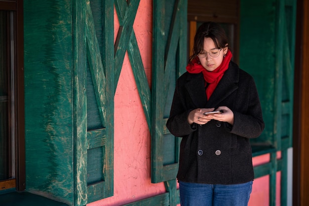 una chica con un teléfono en las manos se encuentra en la calle cerca de una casa rosa con persianas verdes