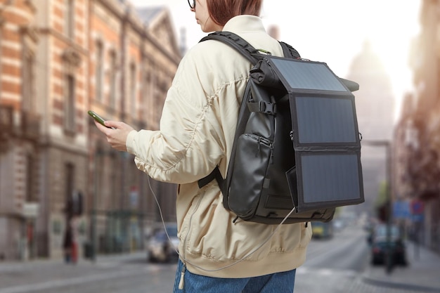 Chica con teléfono móvil en las manos y batería solar portátil en mochila viajando por una ciudad