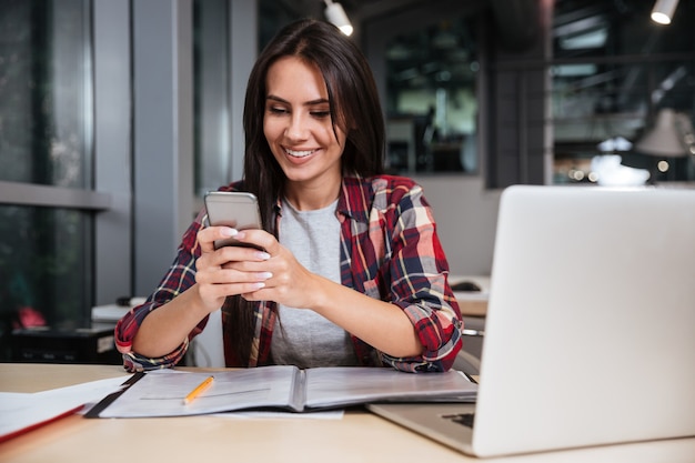 Chica con teléfono sentado junto a la mesa con documentos y portátil en la oficina.