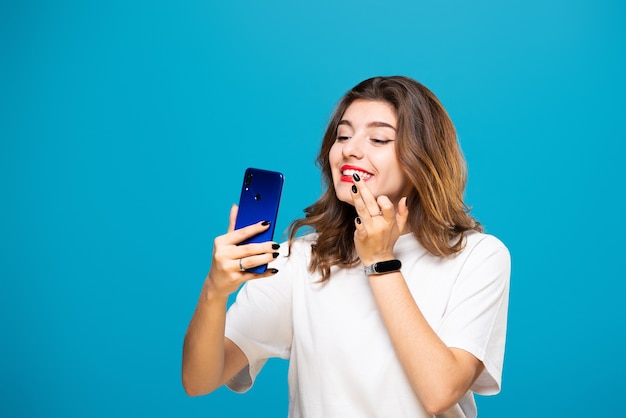 Chica con un teléfono en sus manos sonríe y hace maquillaje, aislado en un azul.