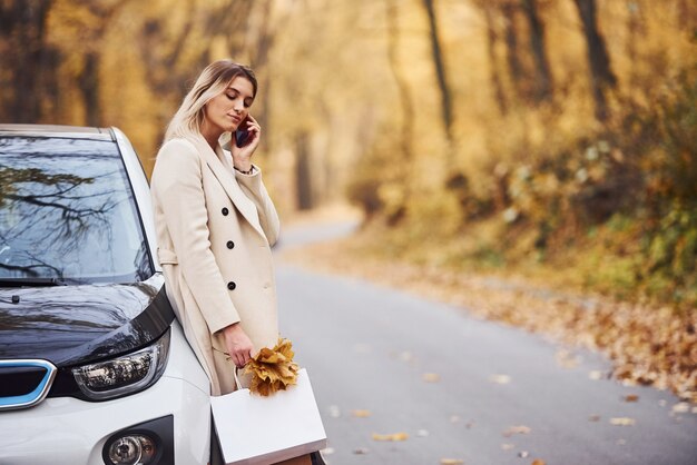 Chica tiene una llamada cuando se para cerca del coche. Automóvil nuevo y moderno en el bosque.