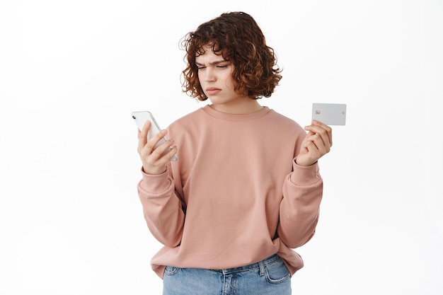 La chica tiene un problema con la aplicación del teléfono móvil. Mujer con tarjeta de crédito y teléfono inteligente, mira fijamente la pantalla con cara confusa y molesta, paga en línea, de pie sobre fondo blanco