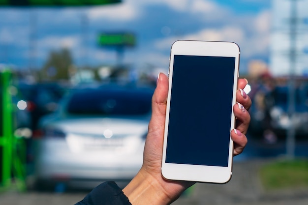 Chica tiene un teléfono inteligente en sus manos. Simulacros de teléfono con pantalla blanca sobre el fondo de un coche eléctrico en una estación de carga.