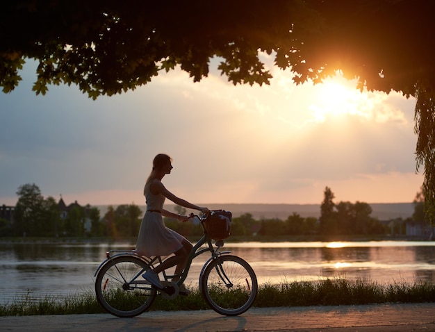 Chica tierna monta una bicicleta retro con cesta en la carretera cerca del río al atardecer