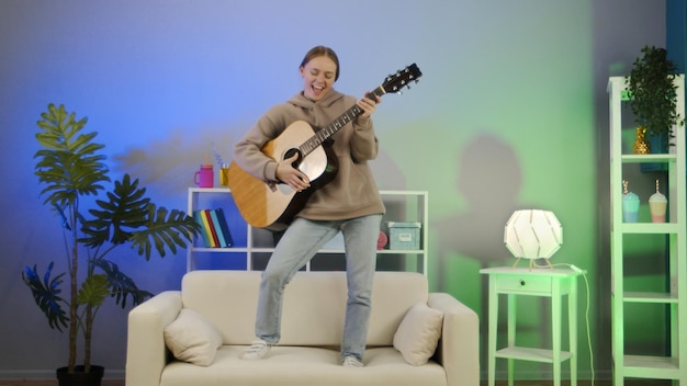 Chica tocando la guitarra acústica en casa Mujer joven divertida tocar un instrumento musical en el sofá