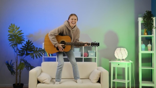 Chica tocando la guitarra acústica en casa Mujer joven divertida tocar un instrumento musical en el sofá