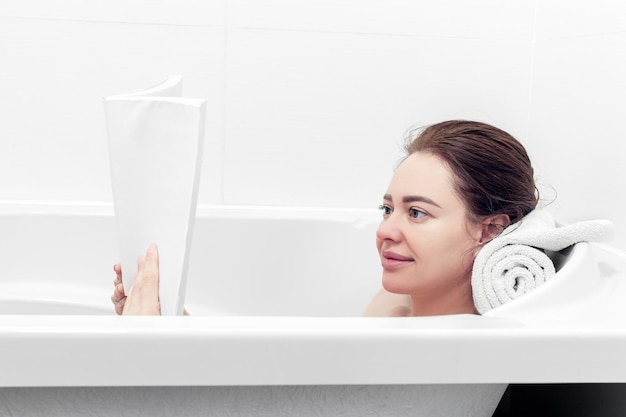 Chica toma un baño mientras lee un libro en un baño blanco