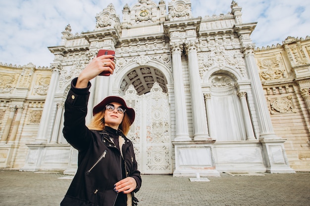 Chica toma una selfie en la plaza frente a Dolmabahce, Estambul, Turquía. Una joven turista con sombrero y abrigo es fotografiada en el teléfono frente al Palacio del Sultán en Estambul.