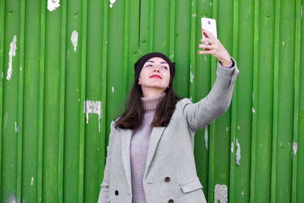 Chica toma una selfie en una puerta industrial verde. con sombrero y abrigo. Lugar para escribir. vestido con una pagoda fría. ropa y estilo. emociones naturales