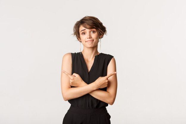 Chica tonta despistada sonriendo y señalando con el dedo hacia los lados, pidiendo ayuda con la elección, tomando decisiones, de pie sobre blanco.