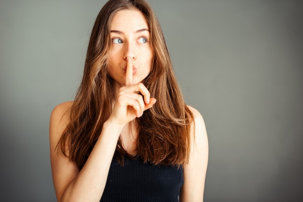 Una chica con un top negro con el pelo está pidiendo paz y tranquilidad, haciendo un gesto con un dedo delante de la boca, diciendo shhhh en una pared gris. Sin maquillaje, sin retoques.