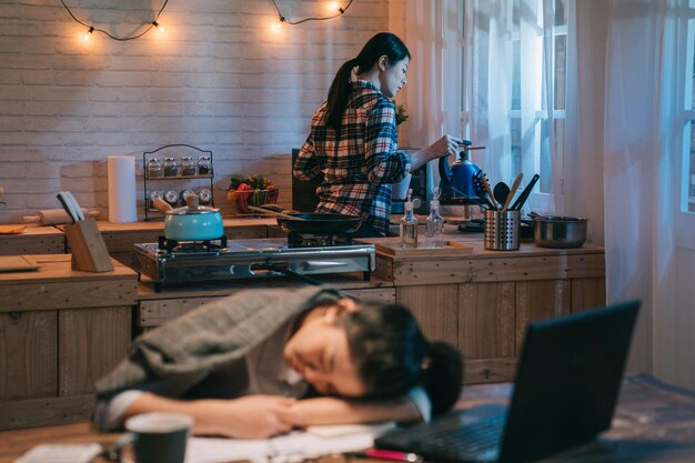 chica trabajadora cansada con papeleo de computadora portátil y café durmiendo en una mesa de madera. una joven trabajadora cubierta con una manta por un compañero de cuarto. Amiga de pie en la cocina de la casa de la noche de fondo