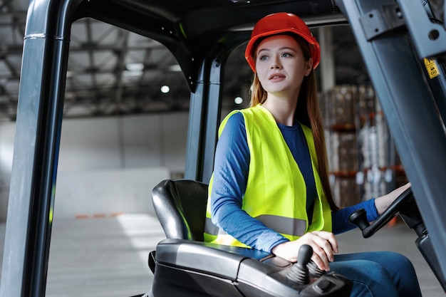 Foto chica trabajadora linda mujer joven con sombrero y ropa de trabajo conduciendo una carretilla elevadora mirando hacia otro lado