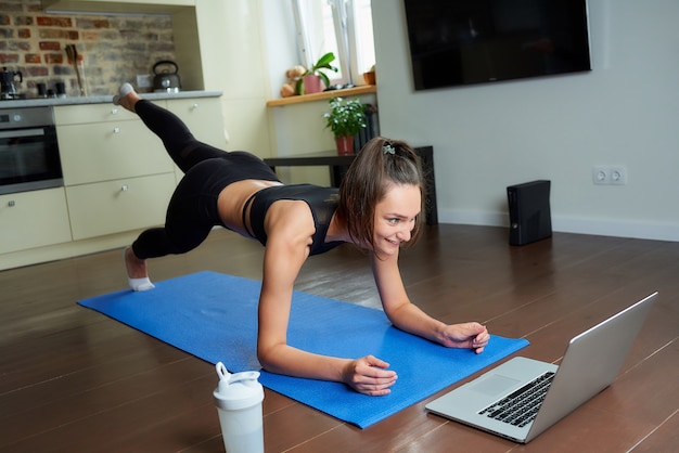 Una chica con un traje ajustado de entrenamiento está haciendo ejercicios de glúteos con un video en línea en una computadora portátil.