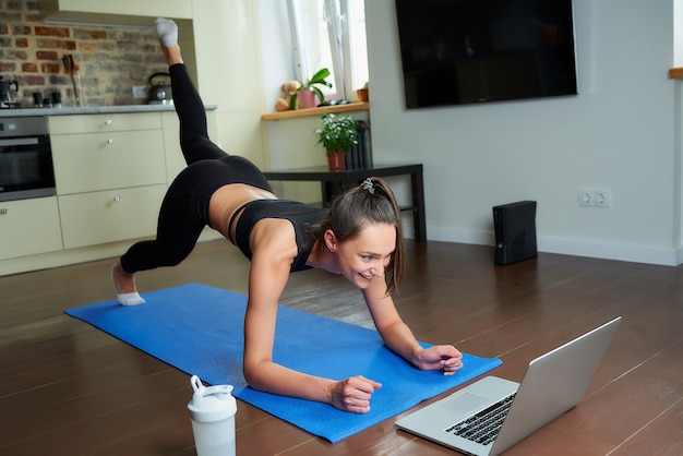 Una chica con un traje ajustado de entrenamiento está haciendo ejercicios de glúteos con un video en línea en una computadora portátil.