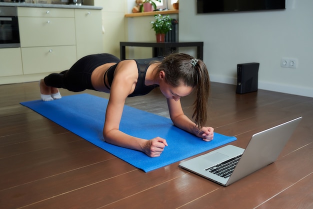 Una chica con un traje ajustado de entrenamiento negro está haciendo plancha mirando un video en línea en una computadora portátil.