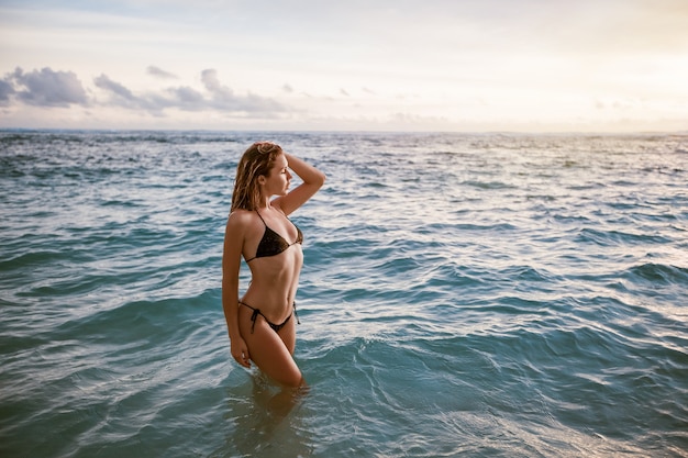 Chica en traje de baño bikini posando en el océano al atardecer