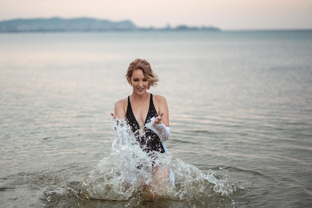 una chica con traje de baño negro y camisa blanca en el mar se ríe y salpica