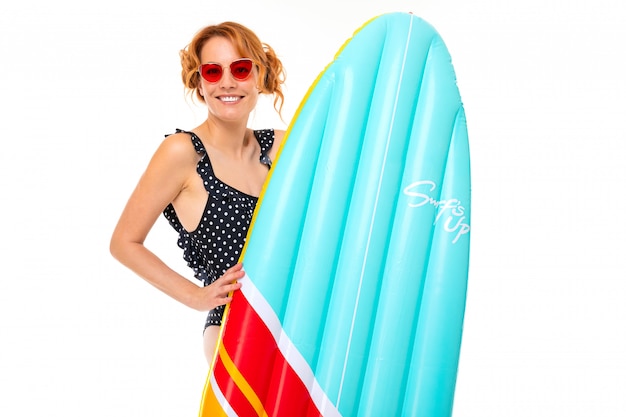 Chica en traje de baño retro y gafas con una tabla de surf en una pared blanca