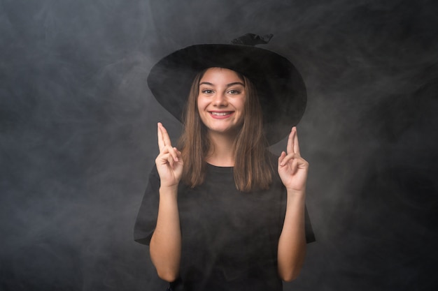 Chica con traje de bruja para fiestas de halloween sobre pared oscura aislada con dedos cruzando