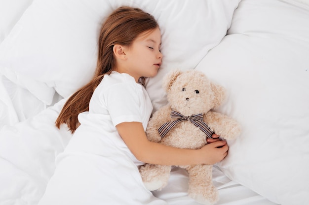 Chica tranquila y agradable abrazando un juguete esponjoso mientras toma una siesta en una cama blanca y cómoda