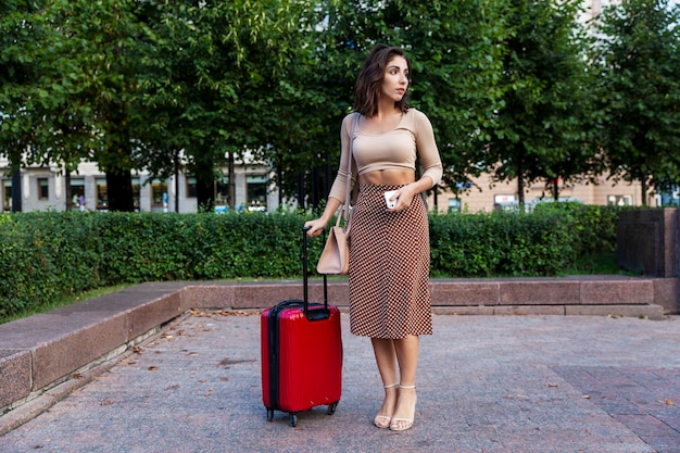 Chica triste y confundida con una maleta en un parque de la ciudad Hermosa morena elegante con falda Viajando sola