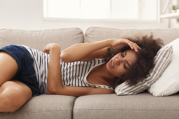 Chica triste con dolor de cabeza. Joven mujer afroamericana sintiendo dolor, acostado en el sofá en casa, espacio de copia