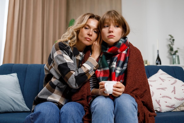 Chica triste con gripe en una manta y con una taza de té y su madre sentada en el sofá