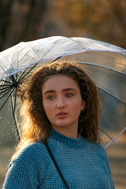 Chica triste con ojos azules y cabello rizado bajo un paraguas transparente Retrato de mujer joven con suéter azul