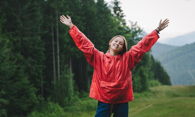 Chica turista alegre de pie en las montañas en clima lluvioso contra el hermoso paisaje de fondo