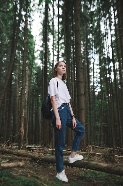 Chica turista en camiseta blanca con una mochila posando para la foto en el bosque de abetos